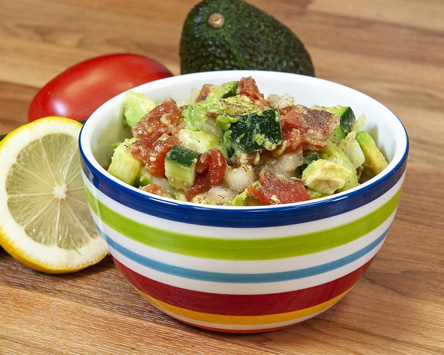 A bowl of diced cucumber, tomato and avocado salad with a lemon and whole tomato beside it