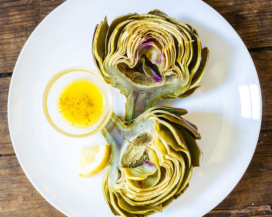 A steamed artichoke on a white plate with a small dish of butter and lemon